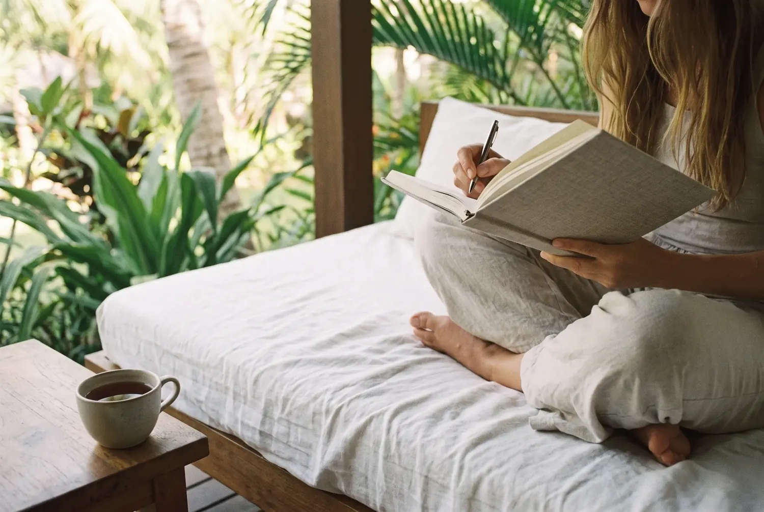 Hands writing in a journal on a daybed surrounded by tropical plants